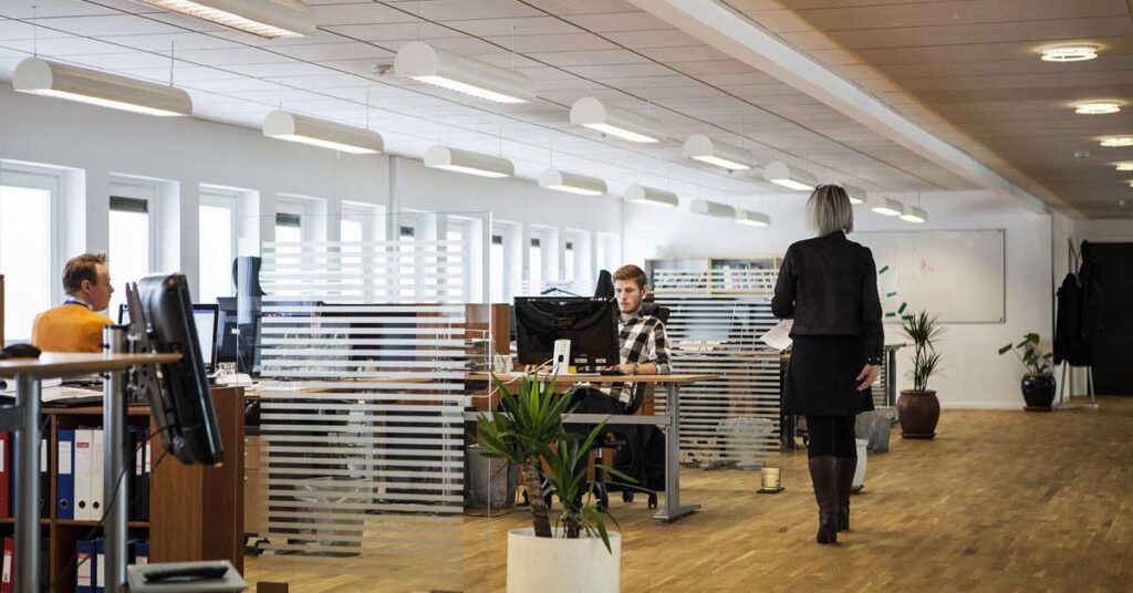 A woman walks through a modern office space featuring a desk and chairs, representing a digital workplace environment. Photo: louisehoffmann83 from Pixabay
